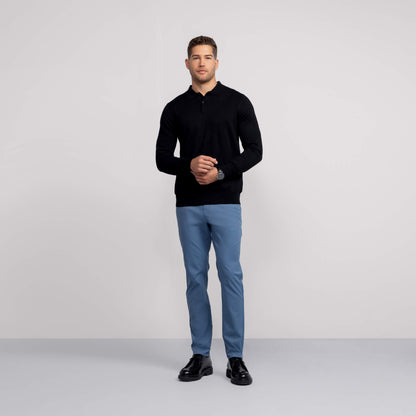 A man stands against a plain, light gray background wearing a black long-sleeve shirt, black shoes, and light blue Jetsetter Tech Pant Slim Fit, with his hands loosely clasped in front of him.