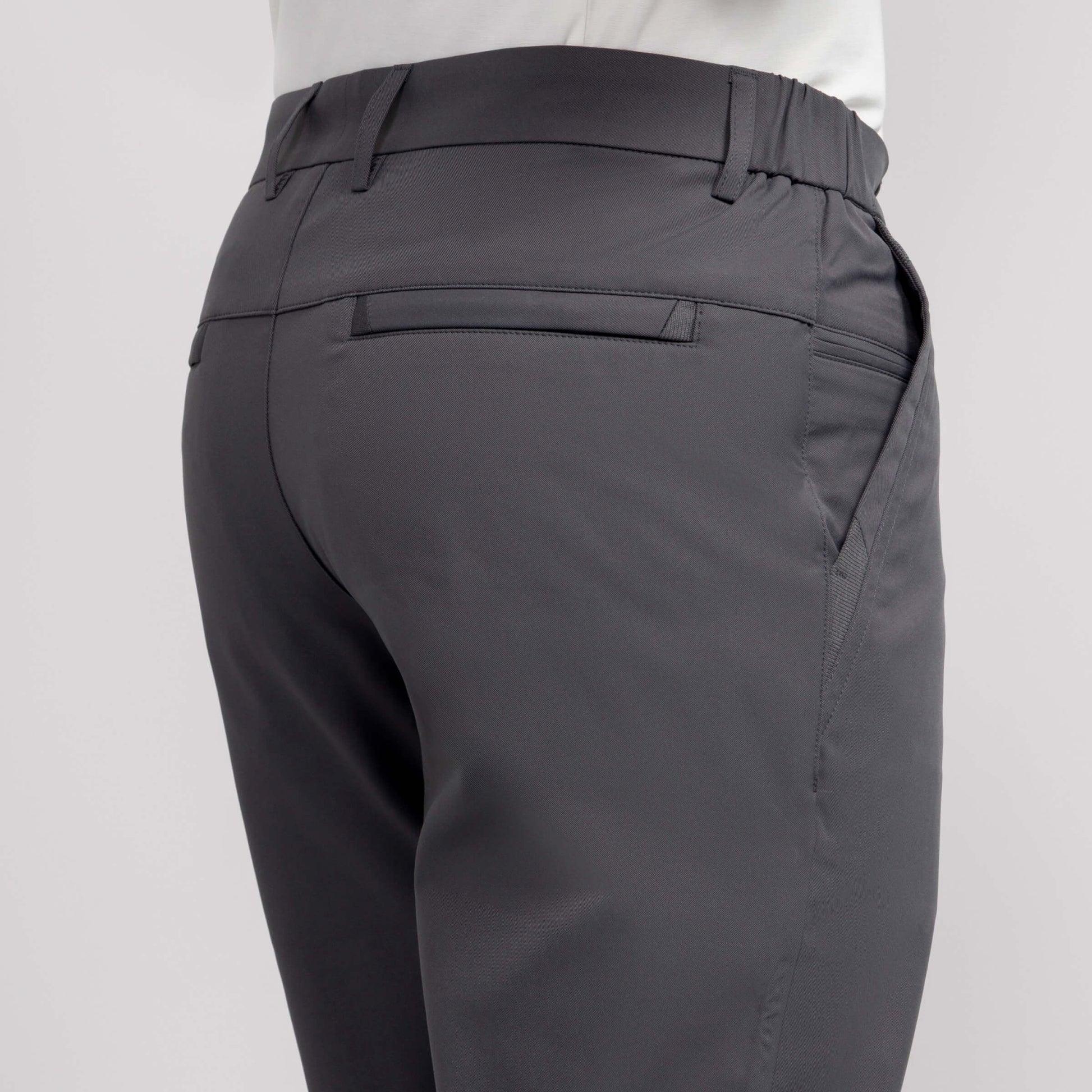 A close-up of the back and side of a person wearing Jetsetter Tech Pant Slim Fit in dark gray with wrinkle-free fabric, featuring a waistband, welt back pocket, and side pocket, paired with a white shirt against a plain background.