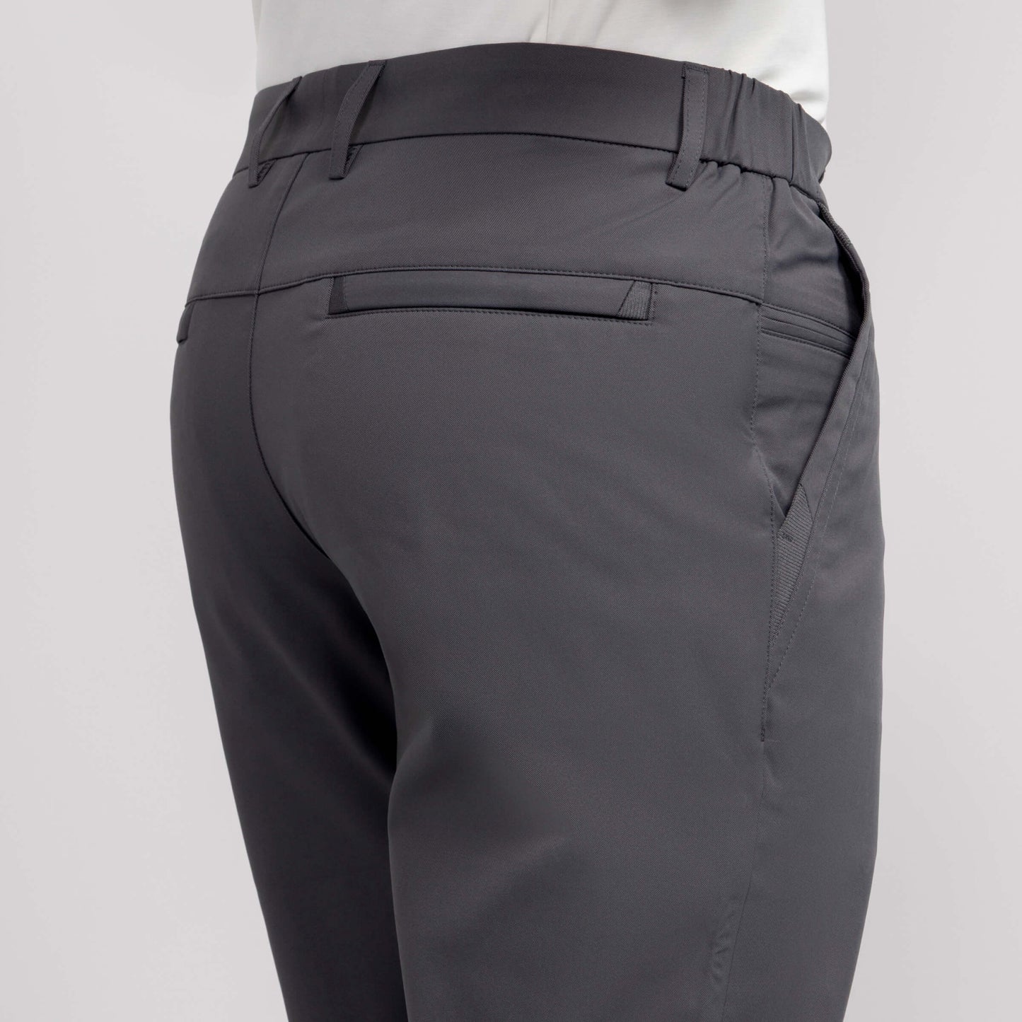 A close-up of the back and side of a person wearing Jetsetter Tech Pant Slim Fit in dark gray with wrinkle-free fabric, featuring a waistband, welt back pocket, and side pocket, paired with a white shirt against a plain background.